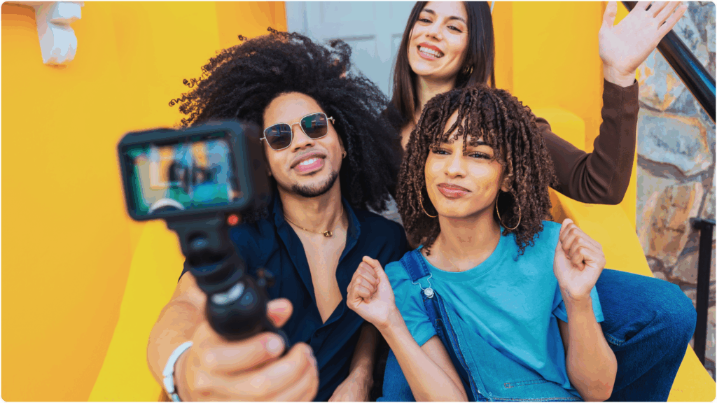 Three young adults are happily posing for a selfie video using a handheld action camera. They are standing against a bright yellow wall, exuding a lively and cheerful vibe. The person in front, wearing sunglasses and a dark shirt, is holding the camera. A woman in a blue shirt with curly hair smiles beside him, while another woman with straight hair in a dark top stands behind them, waving. The scene suggests friendship, social media content creation, or vlogging.